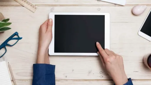 A light coloured table top, from left to right there is a green plant, blue glasses, hands holding a tablet, a phone, a mug and a pen.